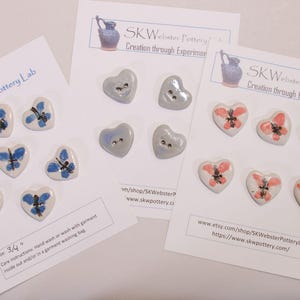 May include: Heart-shaped ceramic buttons with butterfly designs in blue, gray, and coral colors. The buttons are attached to white cards with the text "SK Webster Pottery Lab" and care instructions. The buttons are approximately 3/4 inch in size.
