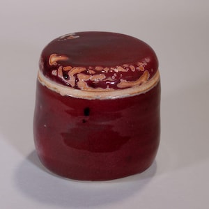 Puede incluir: Un pequeño tarro cilíndrico de cerámica con tapa. El tarro es de color burdeos intenso con un acabado brillante. La tapa tiene un diseño más claro y texturizado alrededor del borde. El tarro es probablemente para almacenamiento o fines decorativos.