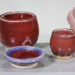 May include: A set of three small, red-brown ceramic pieces: a cup, a bowl, and a saucer. The cup has a handle and the saucer has a purple rim.