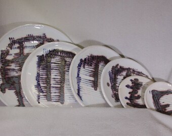May include: Set of five white ceramic plates with purple and grey abstract designs. The plates are nested together in size order, with the largest plate at the bottom and the smallest plate at the top.