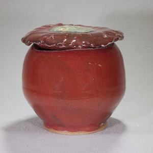 Pode incluir: Um pote de cerâmica vermelho com uma tampa em forma de flor. A tampa tem um esmalte verde e branco.