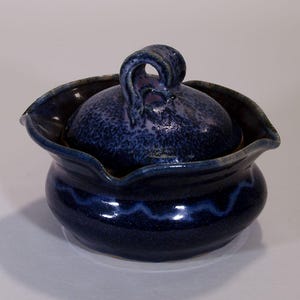 Könnte beinhalten: Ein handgefertigter Keramiktopf mit Deckel, mit einer tiefblauen Glasur mit gesprenkelten Details. Der Topf hat einen wellenförmigen Rand und einen dekorativen Griff auf dem Deckel. Ein blaues Wellenmuster umgibt den Boden.
