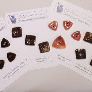 May include: An assortment of handmade ceramic buttons in various shapes and colors. The buttons include dark brown triangles, square, and triangular buttons with a reddish-brown hue. The text "SKW Webster Pottery Lab" is visible.
