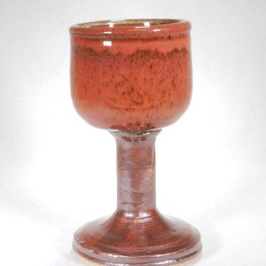 May include: A red ceramic goblet with a wide base and a tall, slender stem. The goblet has a speckled glaze and a slightly uneven rim.