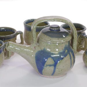 May include: A ceramic tea set consisting of a teapot with a high arched handle, three mugs with handles, and a lidded sugar bowl. Each piece features a speckled light green glaze with deep blue drip accents around the rims and bodies.