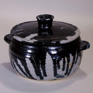 Puede incluir: Una olla de cerámica con tapa, con un diseño de esmalte negro y blanco. La olla tiene dos asas pequeñas y un pomo en la tapa. La olla mide aproximadamente 15 cm de diámetro y 13 cm de altura.