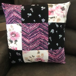May include: A square throw pillow featuring a patchwork design of nine alternating fabric squares. The pattern includes a purple and black chevron print, a black fabric with small light blue floral accents, and a white fabric with large pink flowers.