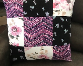 May include: A square throw cushion featuring a patchwork design of nine alternating fabric squares. The pattern includes a purple and black chevron print, a black fabric with small light blue floral accents, and a white fabric with large pink flowers.