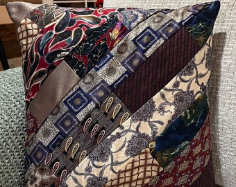 May include: A square throw pillow constructed from diagonal strips of various silk neckties. The fabric features a mix of patterns including paisley, geometric squares, and lattice designs in a color palette of burgundy, navy blue, cream, and brown.