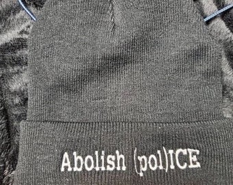May include: Black knit beanie with white embroidered text that reads "Abolish (pol)ICE".