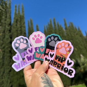 May include: A hand holds a collection of colorful stickers with paw print designs. Each sticker features a paw print, the words "FIP Warrior," and a heart. The stickers have a holographic effect and are set against a blue sky.