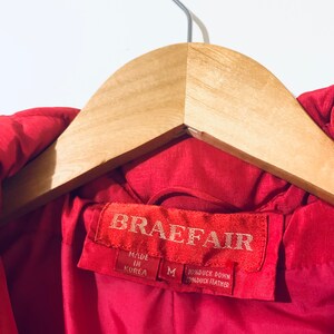May include: Red down jacket with a label that reads "Braefair" and "Made in Korea". The label also indicates the jacket is size M and is made with 80% duck down and 20% duck feather.