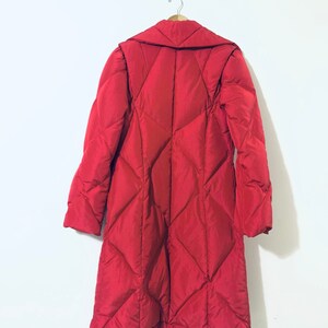 May include: A long red quilted coat with a large collar. The coat is hanging on a hanger against a white wall.
