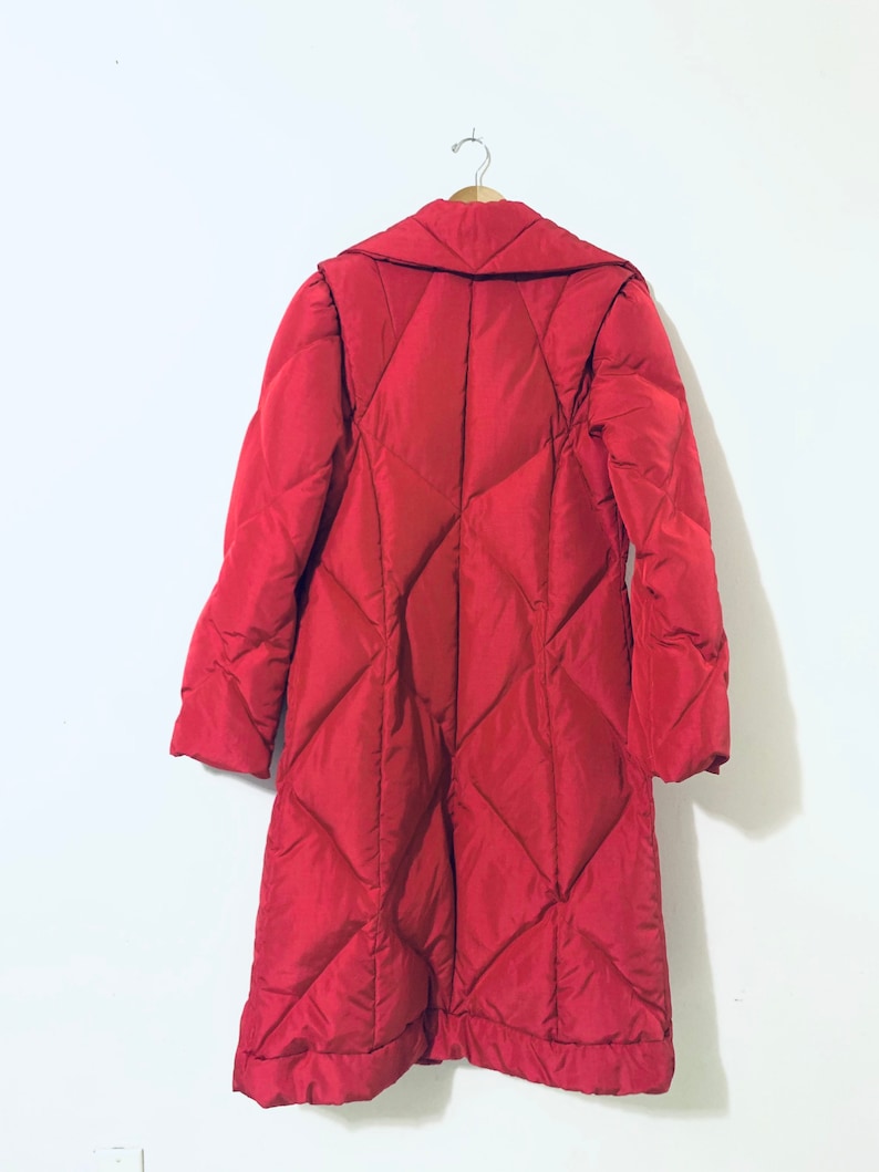 May include: A long red quilted coat with a large collar. The coat is hanging on a hanger against a white wall.