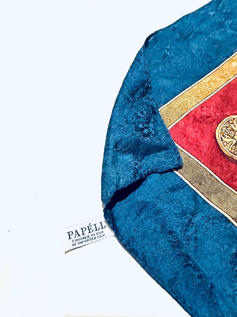 Puede incluir: Un pa&ntilde;uelo de seda azul con un borde estampado en oro y rojo. El pa&ntilde;uelo tiene una etiqueta blanca que dice "Papell, Finished in USA of Imported Silk".