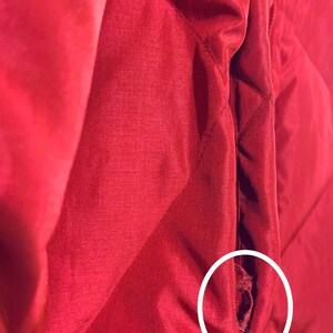 May include: A close-up of a red quilted jacket with a small tear in the seam. The tear is circled in white.