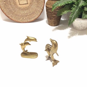 Puede incluir: Dos esculturas de delfines de latón dorado. Los delfines están saltando y parecen estar en el aire. Las esculturas están sobre una superficie blanca.