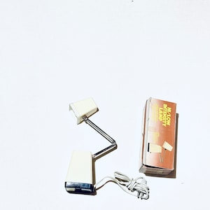 May include: A white desk lamp with a flexible arm and a white shade. The lamp is plugged in and has a switch on the base. The lamp is packaged in a brown box with the text "Hi/Low Intensity Lamp" printed on it.