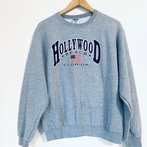 Puede incluir: Sudadera gris con un gr&aacute;fico en azul marino "HOLLYWOOD BEACH FLORIDA" con una bandera estadounidense.