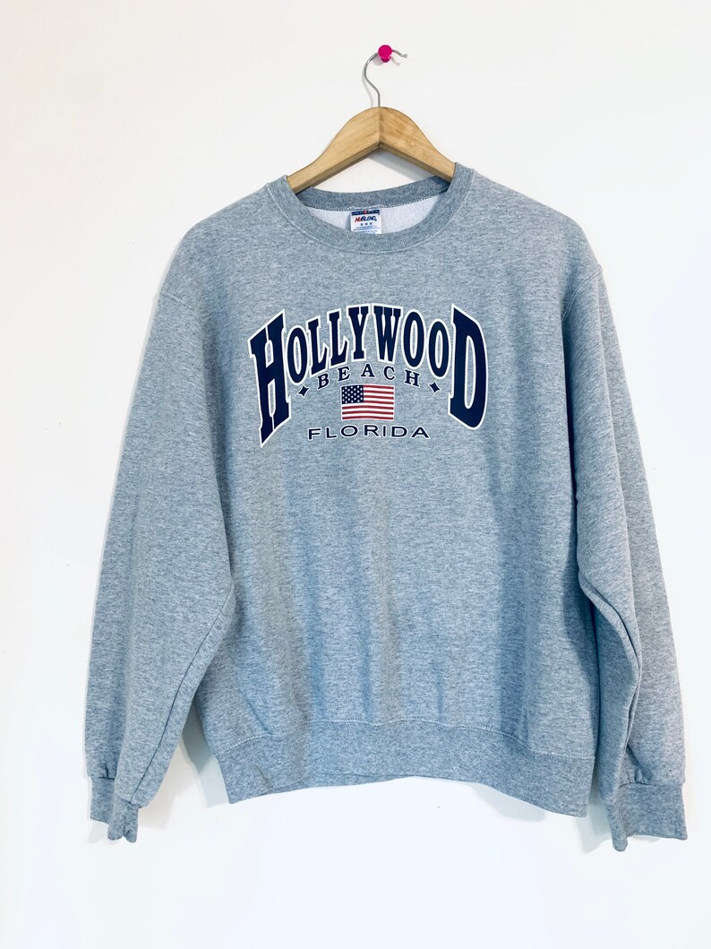 Puede incluir: Sudadera gris con un gr&aacute;fico en azul marino "HOLLYWOOD BEACH FLORIDA" con una bandera estadounidense.