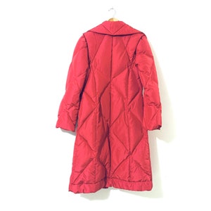 May include: A long red quilted coat with a collar and a button closure. The coat is hanging on a hanger against a white background.