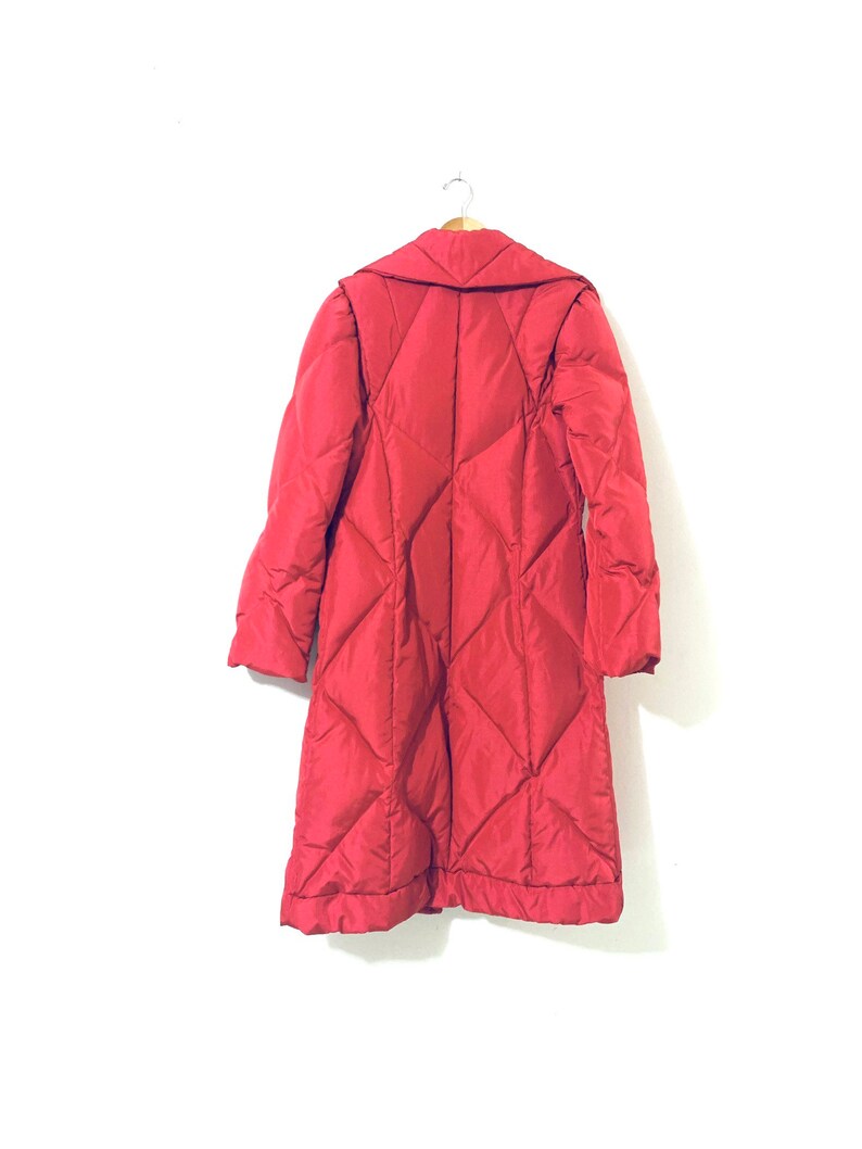 May include: A long red quilted coat with a collar and a button closure. The coat is hanging on a hanger against a white background.