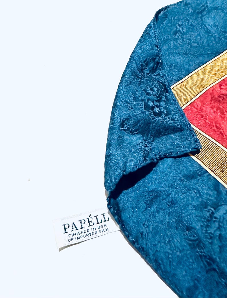 Puede incluir: Un pa&ntilde;uelo de seda azul con un borde estampado en oro y rojo. El pa&ntilde;uelo tiene una etiqueta blanca que dice "Papell, Finished in USA of Imported Silk."