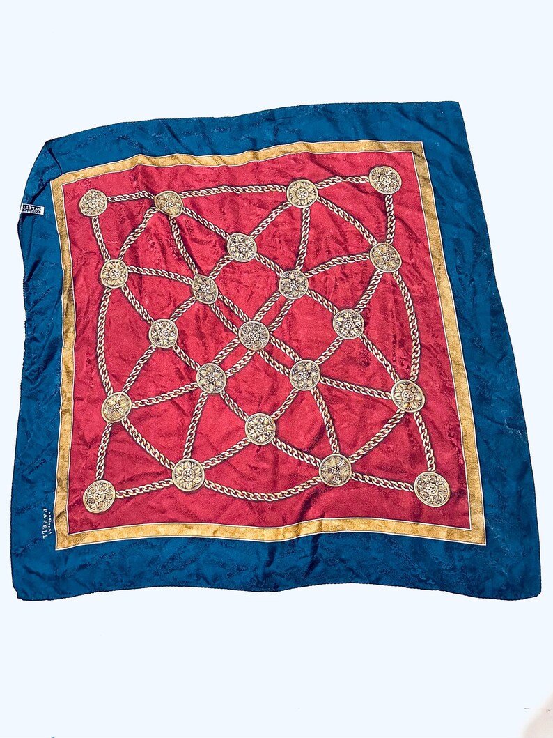 Puede incluir: Un pa&ntilde;uelo de seda rojo y azul con un patr&oacute;n de cadena dorada y medallones. El pa&ntilde;uelo tiene un borde dorado y el texto "PARELL" es visible en la esquina inferior izquierda.