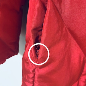 May include: A close-up of a red quilted jacket with a small tear in the fabric near the pocket.