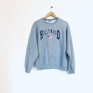 Puede incluir: Sudadera gris con un gr&aacute;fico azul marino de "HOLLYWOOD BEACH FLORIDA" con una peque&ntilde;a bandera estadounidense en el centro.