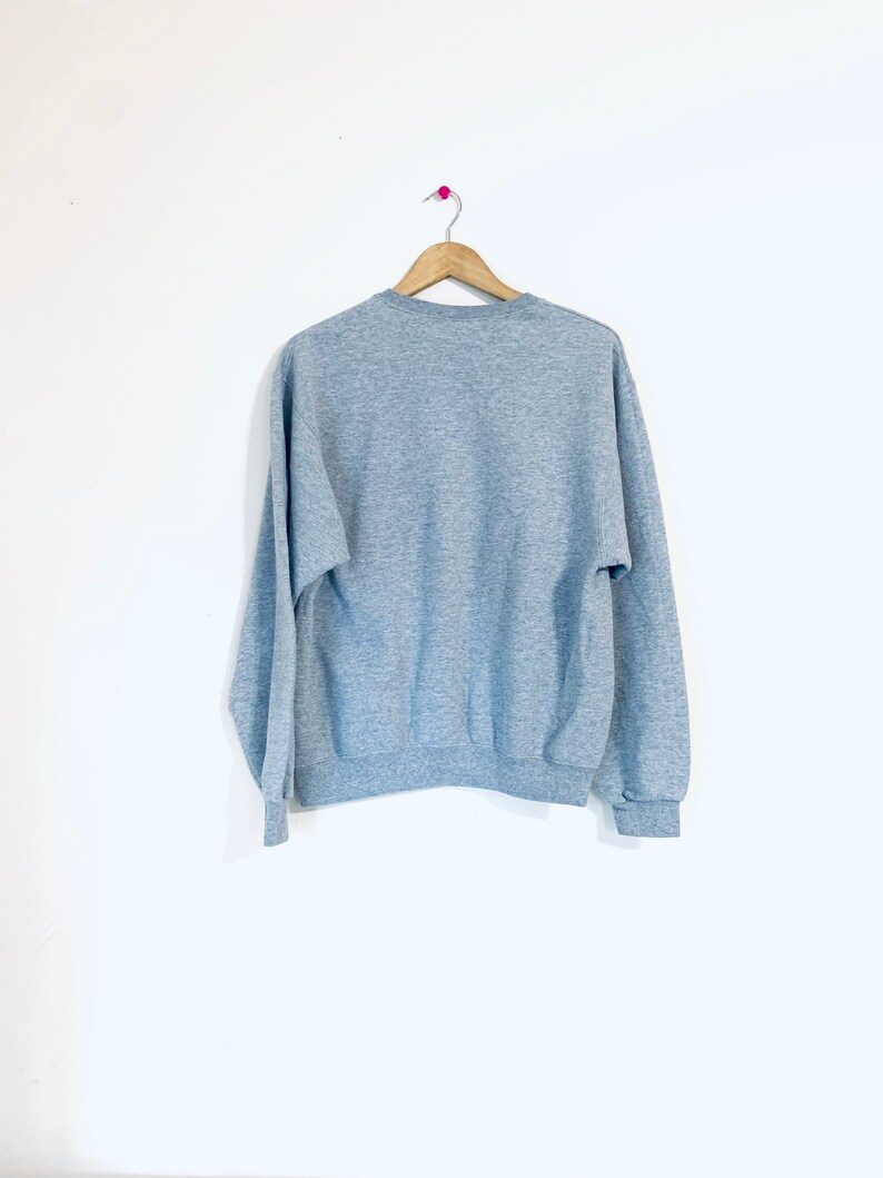 Puede incluir: Una sudadera gris claro colgada de una percha de madera sobre un fondo blanco.