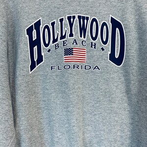 Puede incluir: Sudadera gris con un gr&aacute;fico azul marino que dice "HOLLYWOOD BEACH FLORIDA" con una bandera estadounidense en el centro.