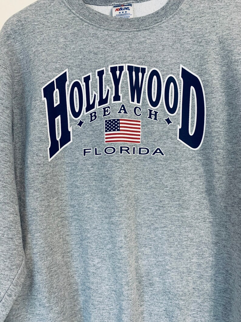 Puede incluir: Sudadera gris con un gr&aacute;fico azul marino que dice "HOLLYWOOD BEACH FLORIDA" con una bandera estadounidense en el centro.