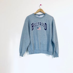 Puede incluir: Sudadera gris con un gr&aacute;fico azul marino y blanco que dice "Hollywood Beach Florida" con una bandera estadounidense.
