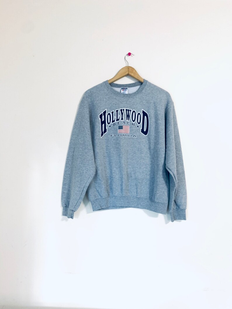 Puede incluir: Sudadera gris con un gr&aacute;fico azul marino y blanco que dice "Hollywood Beach Florida" con una bandera estadounidense.