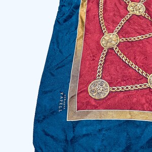 Puede incluir: Un pa&ntilde;uelo de seda azul y rojo con un patr&oacute;n de cadena dorada y medallones dorados. El pa&ntilde;uelo tiene el texto "ADRIANNA PAPE'LL" en la esquina inferior izquierda.