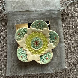 May include: A ceramic flower-shaped coaster with a floral design in shades of green, yellow, and red. The coaster is inside a sheer, light gray drawstring bag, resting on a textured gray surface. The coaster has a cream-colored base.