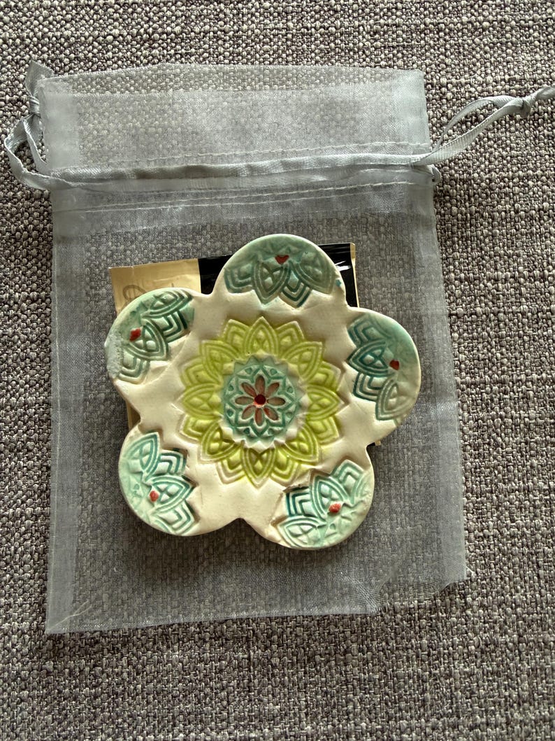 May include: A ceramic flower-shaped coaster with a floral design in shades of green, yellow, and red. The coaster is inside a sheer, light gray drawstring bag, resting on a textured gray surface. The coaster has a cream-colored base.