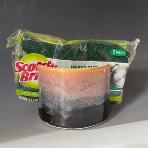 May include: A ceramic mug with a brown, white, and black glaze. The mug is sitting on a white surface next to a package of Scotch-Brite heavy duty scrub sponges. The package says "1 Pack" and "Heavy Duty".