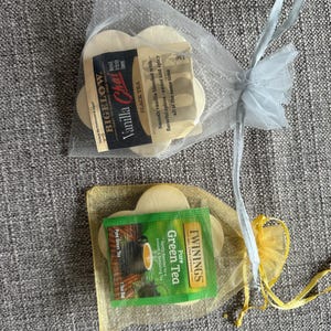May include: Two small gift bags, one silver and one yellow, each containing tea and flower-shaped cookies. One bag has Bigelow Vanilla Chai tea, the other has Twinings Pure Green Tea. The bags are tied with ribbons.