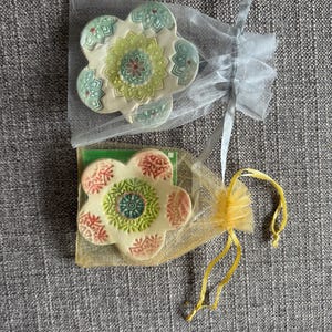 May include: Two flower-shaped ceramic coasters with intricate, colorful designs. One coaster is in a sheer white bag, the other in a yellow bag. The coasters have a cream base with green, red, and blue accents.