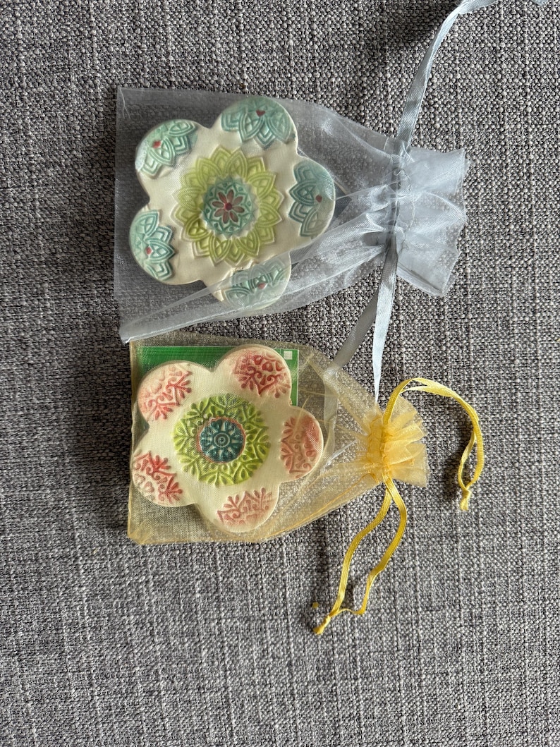 May include: Two flower-shaped ceramic coasters with intricate, colorful designs. One coaster is in a sheer white bag, the other in a yellow bag. The coasters have a cream base with green, red, and blue accents.