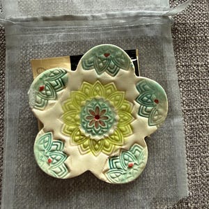 May include: A ceramic flower-shaped coaster with a cream-colored base and green, yellow, and red floral design. The coaster is inside a sheer, drawstring bag, suggesting it's a gift or decorative item.