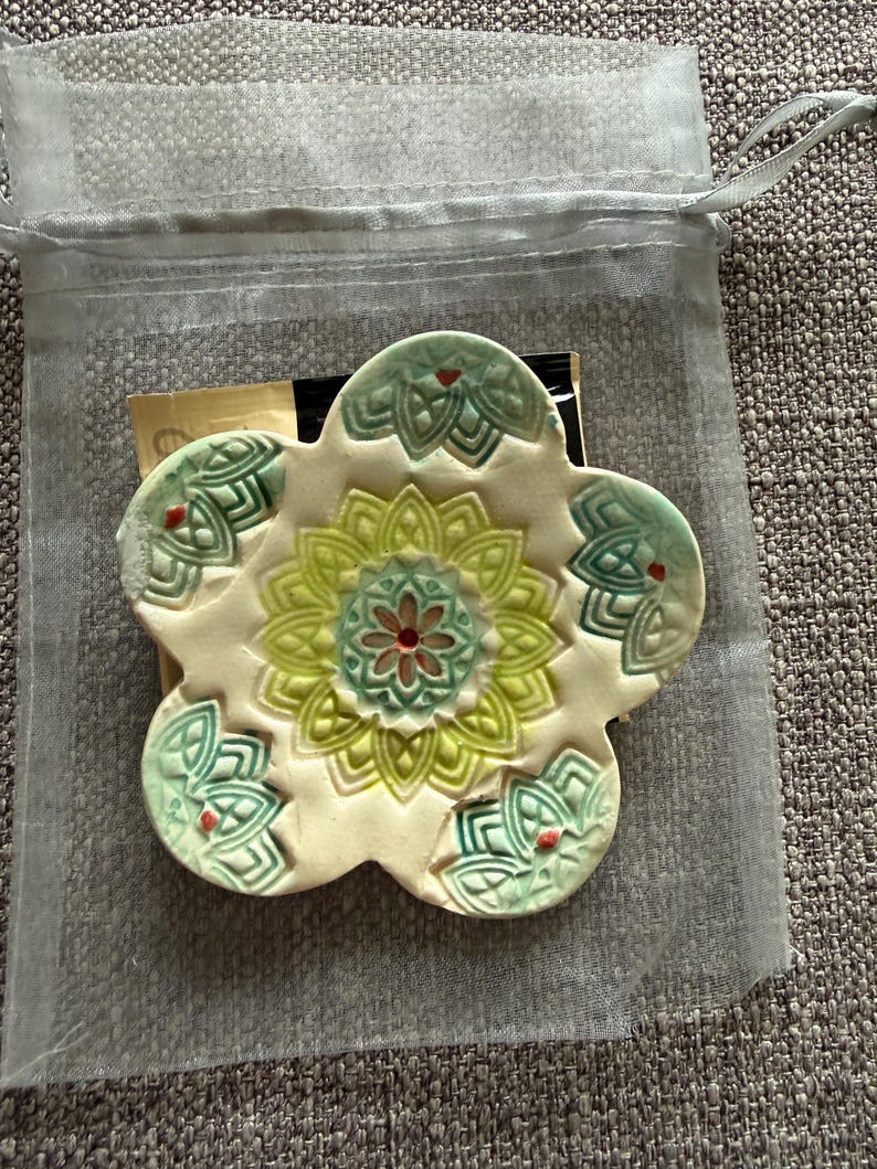 May include: A ceramic flower-shaped coaster with a cream-colored base and green, yellow, and red floral design. The coaster is inside a sheer, drawstring bag, suggesting it's a gift or decorative item.