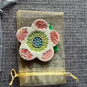 May include: A white ceramic flower-shaped decorative item with red, green, and blue accents. The flower is placed on a small, sheer, beige drawstring bag with a yellow ribbon. The background is a gray textured fabric.