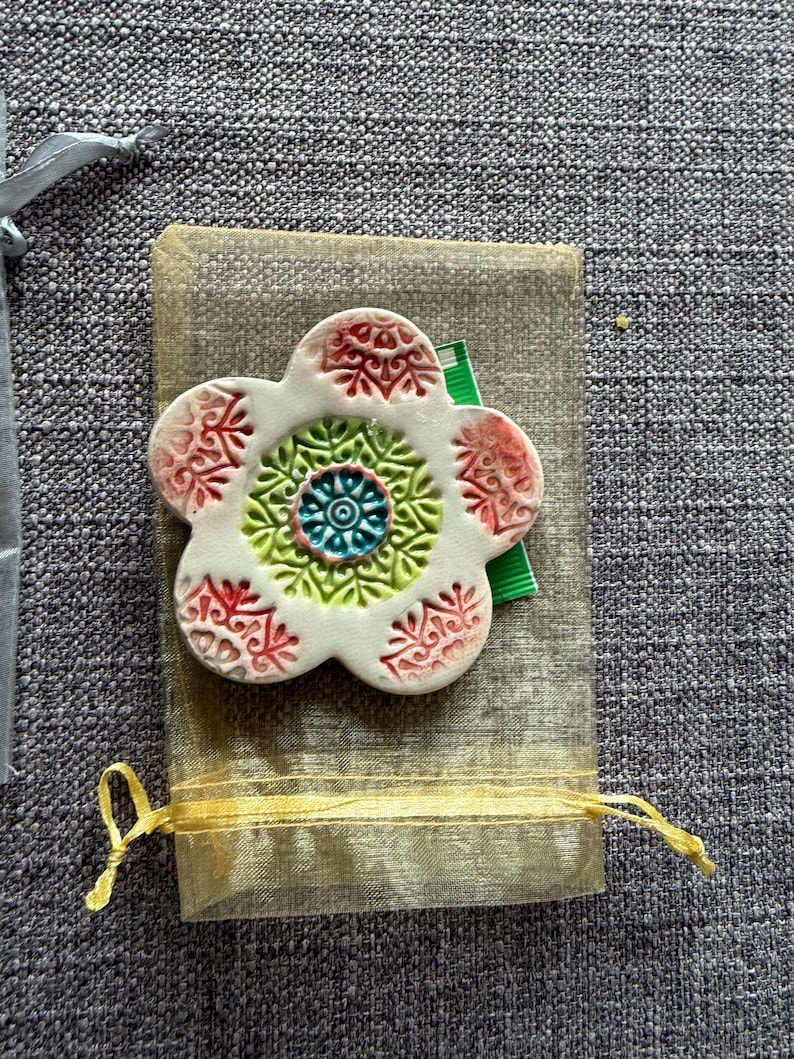 May include: A white ceramic flower-shaped decorative item with red, green, and blue accents. The flower is placed on a small, sheer, beige drawstring bag with a yellow ribbon. The background is a gray textured fabric.