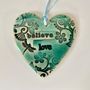 May include: A teal ceramic heart ornament with a black floral design and the words "believe" and "love" in black lettering.