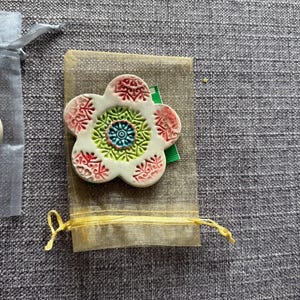 May include: A white ceramic flower-shaped brooch with red, green, and blue decorative patterns. The brooch rests on a small beige fabric pouch tied with a yellow string. A gray fabric background.