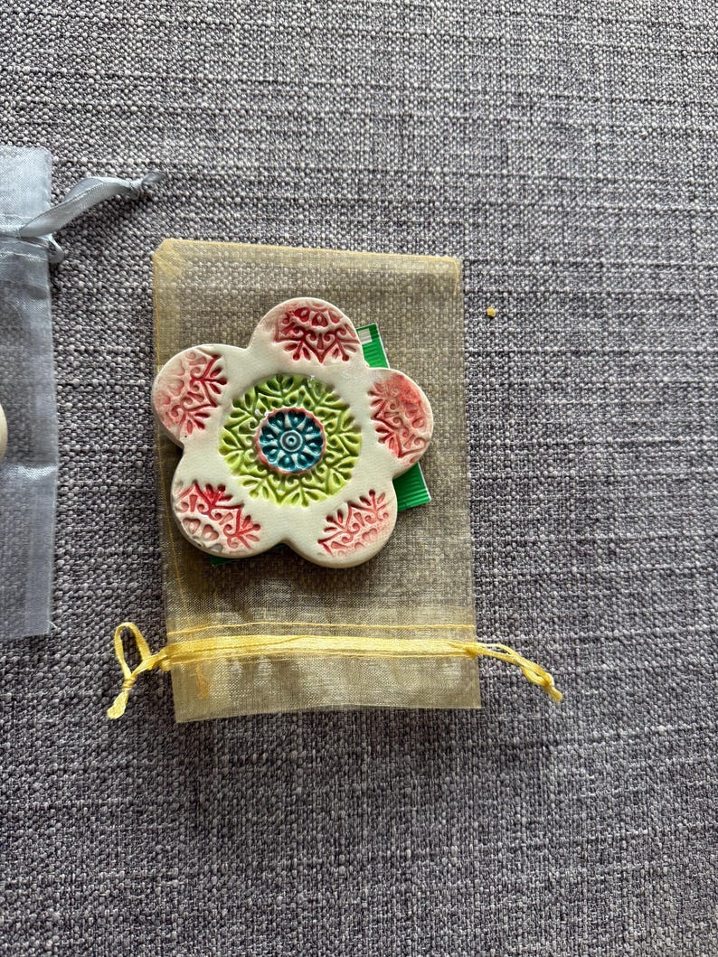 May include: A white ceramic flower-shaped brooch with red, green, and blue decorative patterns. The brooch rests on a small beige fabric pouch tied with a yellow string. A gray fabric background.