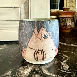 May include: A ceramic mug with a black and white design of a fish. The fish is painted in shades of pink and brown. The mug is sitting on a black surface.